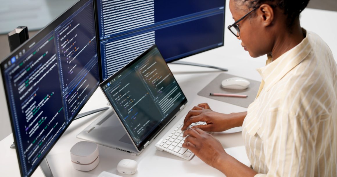A software engineer typing code on a keyboard with multiple monitors displaying programming scripts in a modern office setting.