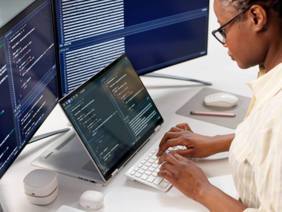 A software engineer typing code on a keyboard with multiple monitors displaying programming scripts in a modern office setting.