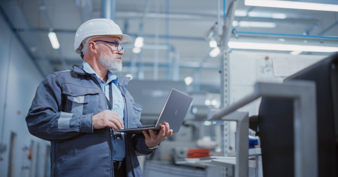 bearded middle-aged engineer in a white hard hat and work suit stands in an industrial facility, holding a laptop and looking upward, representing a heavy industry specialist at work.