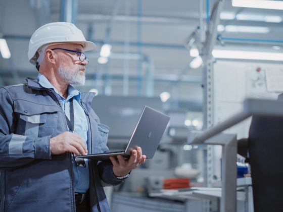 bearded middle-aged engineer in a white hard hat and work suit stands in an industrial facility, holding a laptop and looking upward, representing a heavy industry specialist at work.