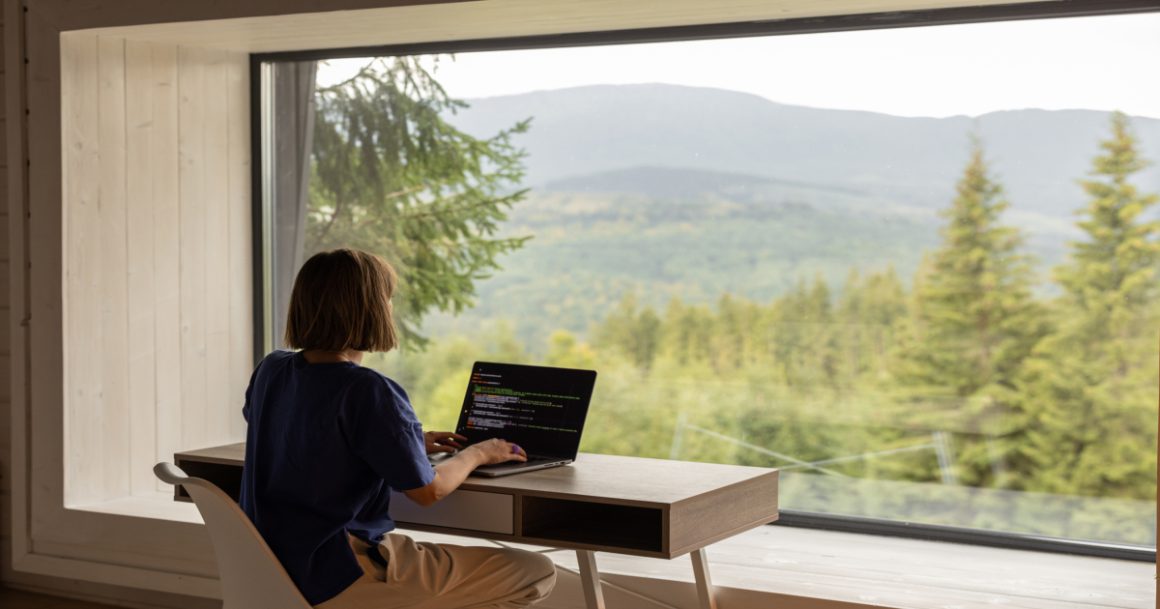 A software engineer working remotely on a laptop with code on screen, seated at a desk in front of a large window overlooking a forested mountain landscape.