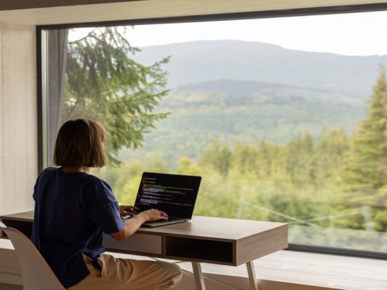 A software engineer working remotely on a laptop with code on screen, seated at a desk in front of a large window overlooking a forested mountain landscape.