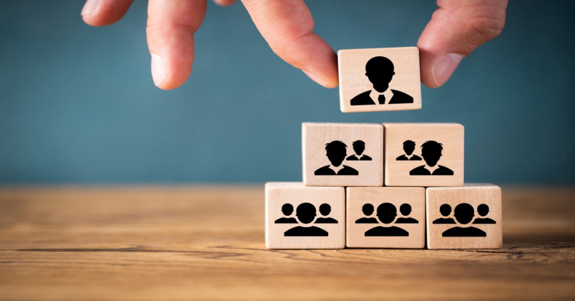 A hand placing a leader block at the top of a pyramid of wooden team blocks, representing leadership qualities and organizational hierarchy