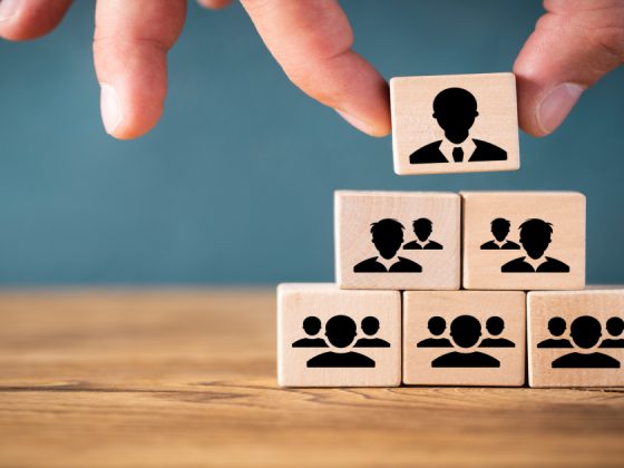 A hand placing a leader block at the top of a pyramid of wooden team blocks, representing leadership qualities and organizational hierarchy