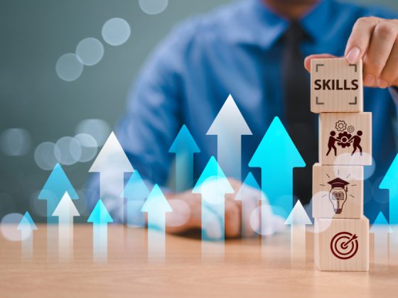 A professional in a blue shirt stacks wooden blocks labeled with icons for skills, teamwork, learning, and goal-setting, surrounded by upward-pointing blue and white arrows symbolizing career growth.