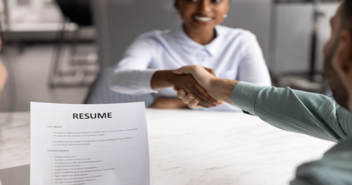 A job candidate and hiring manager shaking hands across a table during an interview, with a resume document visible in the foreground.