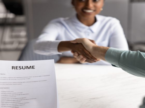 A job candidate and hiring manager shaking hands across a table during an interview, with a resume document visible in the foreground.