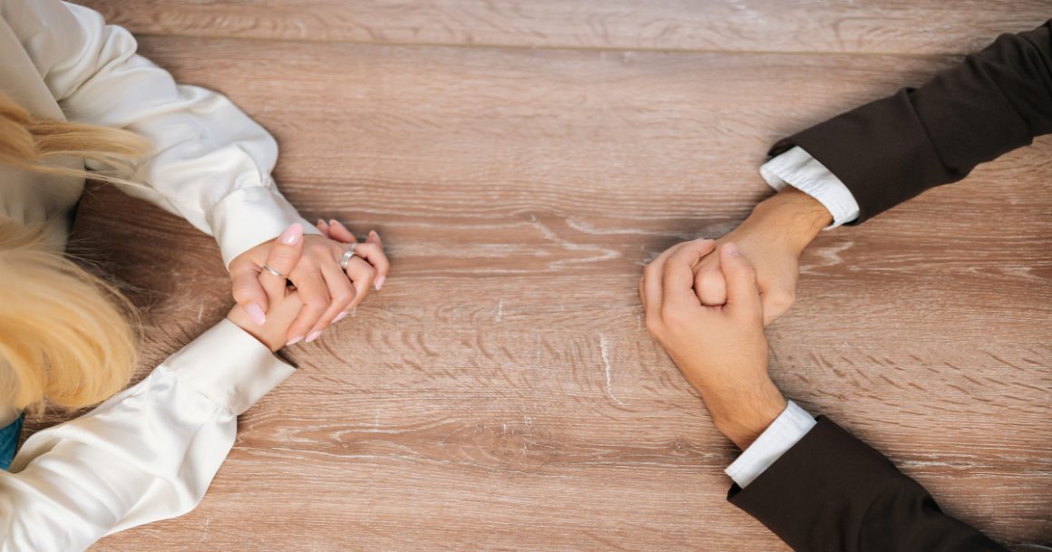 Two people with clasped hands facing each other across a table, illustrating workplace conflict resolution.