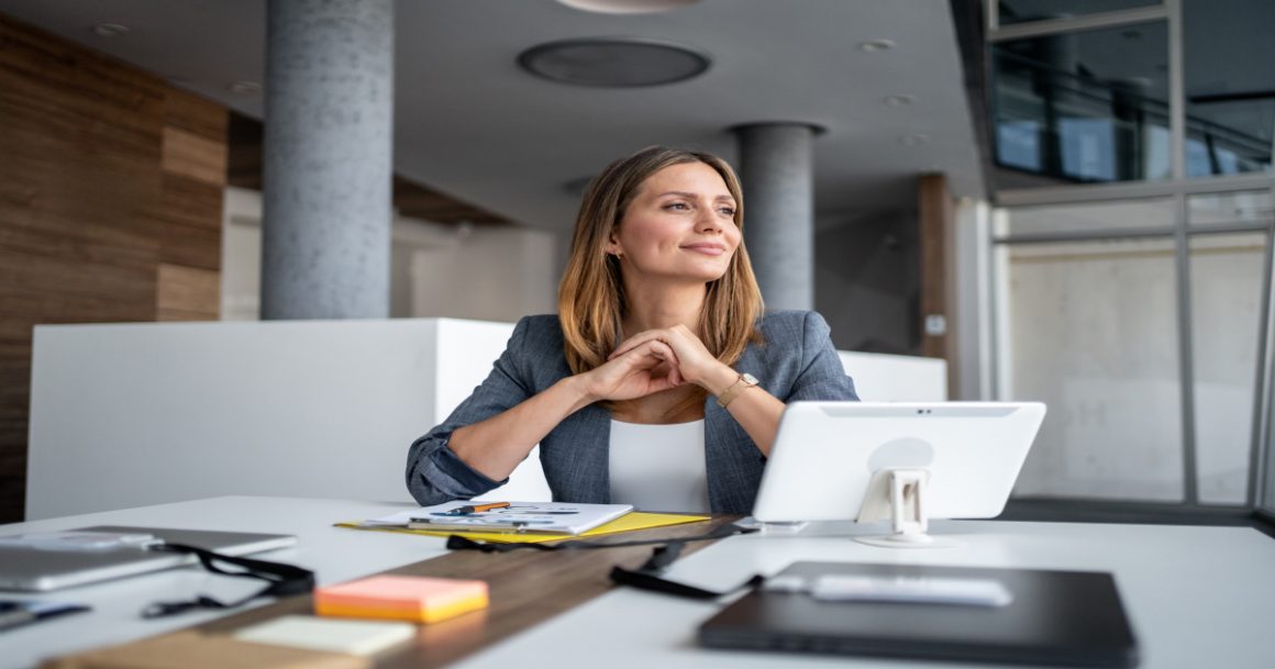 A confident manager sits at her desk with a tablet and notes, smiling thoughtfully while reflecting on her team's goals.