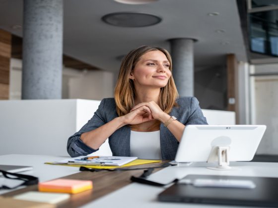 A confident manager sits at her desk with a tablet and notes, smiling thoughtfully while reflecting on her team's goals.