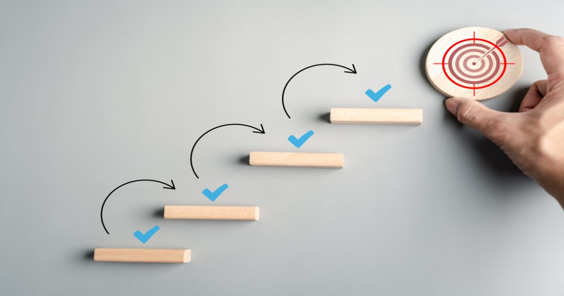Ascending wooden blocks with checkmarks leading to a bullseye, illustrating goal-setting techniques and step-by-step progress.