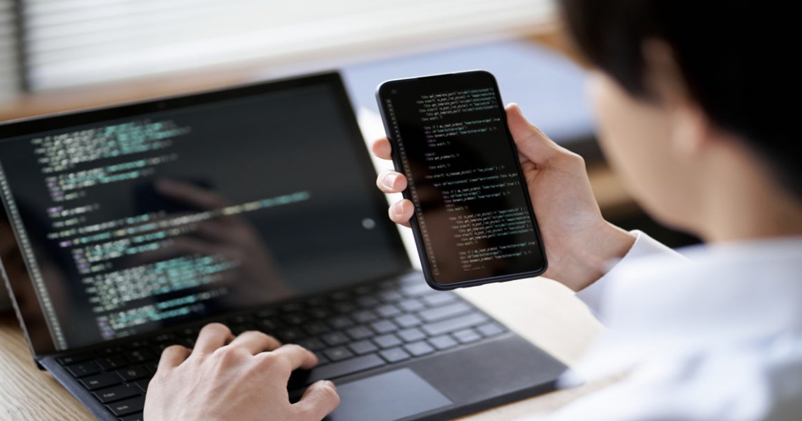 Software tester reviewing code on a smartphone and laptop while testing a mobile app.