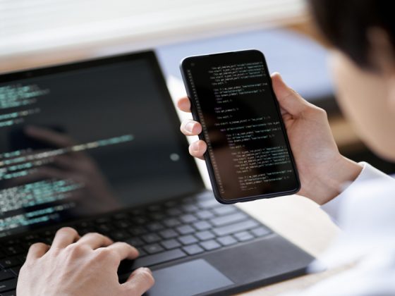 Software tester reviewing code on a smartphone and laptop while testing a mobile app.