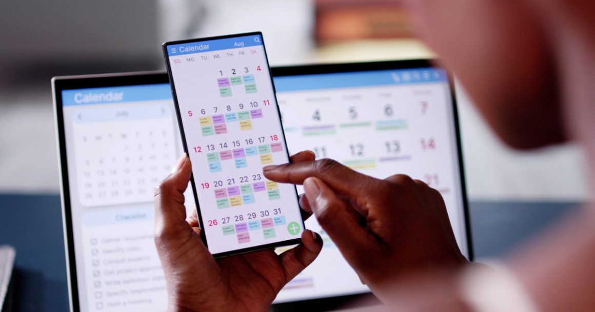 Person reviewing a color-coded work schedule on a smartphone calendar app with a laptop calendar in the background