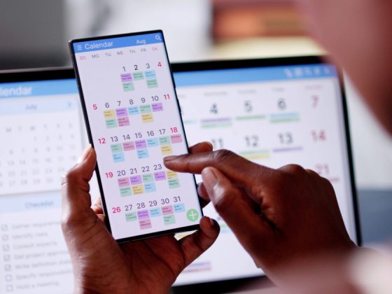 Person reviewing a color-coded work schedule on a smartphone calendar app with a laptop calendar in the background